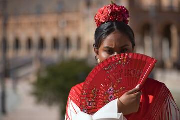 Wall Mural - Portrait of a young black and South American woman in a beige flamenco gypsy costume and red shawl, covering herself from the sun with a fan in Seville in Spain. Concept dance, folklore, flamenco, art