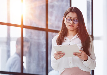 Beautiful confident young busines woman with black hair in a modern office standing near the table with a folder in her hand