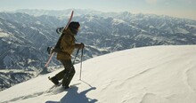 Snowboarder In Snowshoes Climbing Snowy Mountain. Hard Difficult Climb Up High Snowy Winter Mountain With Help Of Snowshoes Ski Poles Athlete Snowboarder Freerider Climbs Mountain Slope With Snowboard