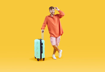 Wall Mural - Studio portrait of happy male tourist. Full body length shot of joyful young man wearing comfortable summer clothes standing with his suitcase on yellow color background. Traveling, holiday concept