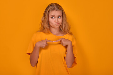 Young shy cute Caucasian woman teenager looks awkwardly to side feeling awkward and puts fingers together in front of chest dressed in casual yellow shirt stands on plain yellow background.