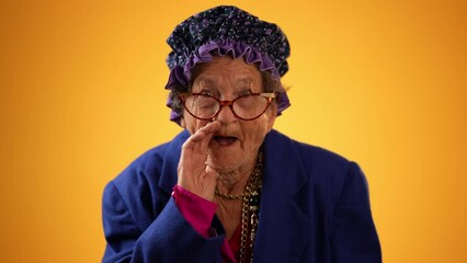 Poster - Funny portrait of smiling happy crazy toothless grandmother with wrinkled skin puts hand to mouth to tell a secret isolated on yellow background studio