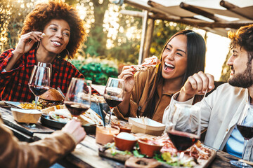 Multiracial friends having fun at barbecue dinner party in garden restaurant - Millennial people cheering red wine sitting at outside bar table - Youth lifestyle, food and beverage concept