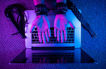 Wall Mural - female sexy hands in bdsm handcuffs with laptop in neon light on dark background