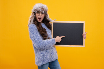 Teenage girl kid hold in warm hat hold blackboard chalkboard with copy space on yellow background. Angry teenager girl, upset and unhappy negative emotion.