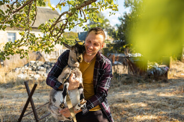 Wall Mural - a happy man of Caucasian appearance, a farmer holds and hugs his favorite goat. a small goat. The concept of a summer countryside. Close to outdoor recreation.