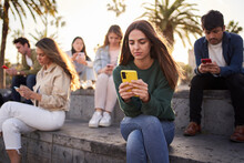Young Caucasian Girl Sitting Concentrated Typing On Mobile With Group Friends In Background. Serious Beautiful Woman Addicted To Technologies Using Phone Outdoors. Social Isolation New Generations.