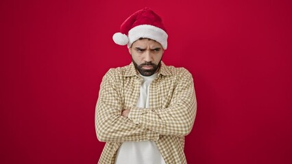 Sticker - Young hispanic man wearing christmas hat standing with arms crossed gesture and unhappy expression over isolated red background