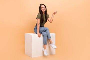 Poster - Full length photo of cute shiny girl dressed khaki t-shirt sitting white cube pointing empty space isolated beige color background