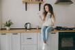 © ViDi Studio - Young housewife woman of African American ethnicity wear casual clothes sweater hold cup drink coffee use mobile cell phone sit on table in light kitchen at home alone. Lifestyle cooking food concept.