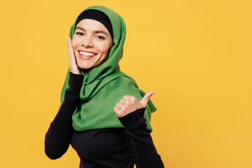 Wall Mural - Side view young arabian muslim woman wear green hijab abaya black clothes point thumb finger on area hold face isolated on plain yellow background. People uae middle eastern islam religious concept.