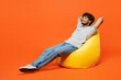 © ViDi Studio - Full bodyside view young smiling happy Indian man he wear t-shirt casual clothes sit in bag chair hold hands behind head neck isolated on orange red color background studio portrait Lifestyle concept