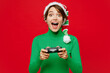 © ViDi Studio - Young gambling woman wear green turtleneck Santa hat posing hold in hand play pc game with joystick console isolated on plain red background. Happy New Year 2024 celebration Christmas holiday concept.