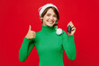 © ViDi Studio - Young amazed woman wear green turtleneck Santa hat posing hold car key fob keyless system showing thumb up isolated on plain red background. Happy New Year 2024 celebration Christmas holiday concept.