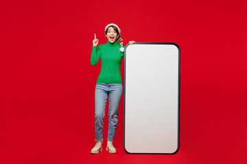 Wall Mural - Full body young woman in green turtleneck Santa hat posing big huge blank screen area mobile cell phone point finger up isolated on plain red background Happy New Year 2024 Christmas holiday concept