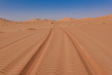 Fototapeta Miasto - Tyre tracks on red sand in the Rub al Khali desert in the background the dunes and a blue sky. Oman.