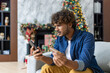 © Liubomir - Cheated and upset Christmas man in living room near decorated Christmas tree, rejected and wrong money transfer, hispanic man holding bank credit card and phone.