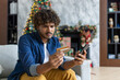 © Liubomir - Cheated and upset Christmas man in living room near decorated Christmas tree, rejected and wrong money transfer, hispanic man holding bank credit card and phone.
