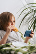 © Konstiantyn Zap - A 9-10-year-old boy with long hair lounges in bed, munching on toast, engrossed in his mobile phone, navigating the internet, and gaming. Capturing the essence of relaxation and idleness