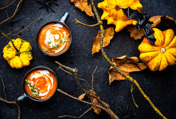 Wall Mural - Festive creamy pumpkin soup with and .thyme. Autumn vegetarian food. Soup mug on black background with fallen oak leaves, pumpkins, spiders, twisted branches and bats. Top view