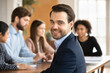 © fizkes - Cheerful successful young businessman posing for camera, smiling with diverse colleagues working in blurred background, talking, brainstorming at meeting table. Business portrait