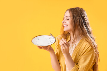 Beautiful young woman with empty plate and spoon on yellow background