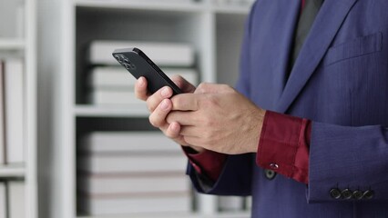 A businessman holding a smartphone is using the mobile phone to send messages online and is typing.
