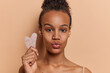 Photo of dark skinned lovely woman holds gua sha stone for face massaging takes care of skin keeps lips rounded looks directly at camera poses over brown background. Morning face care routine concept