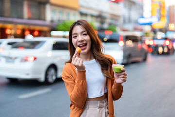 Wall Mural - Young Asian tourists standing and eating Thai desserts. Young beautiful tourists in Chinatown street food market, Bangkok, Thailand