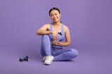 Fototapeta Panele - happy asian woman holding bottle with water, sitting on floor over purple studio background