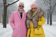 © Glittering Humanity - An eccentric older couple stands in a snowy park, their funky winter coats exuding a vibrant fashion sense as they smile amidst the wintry trees