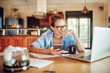 © Geber86 - Happy young woman looking at a smartphone while going over paperwork in the kitchen