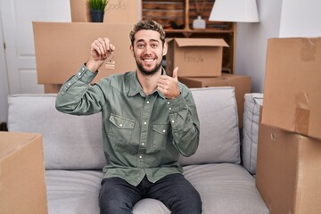 Wall Mural - Hispanic man with beard holding keys of new home smiling happy and positive, thumb up doing excellent and approval sign