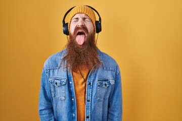 Wall Mural - Caucasian man with long beard listening to music using headphones sticking tongue out happy with funny expression. emotion concept.