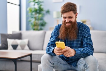 Wall Mural - Young redhead man using smartphone sitting on sofa at home