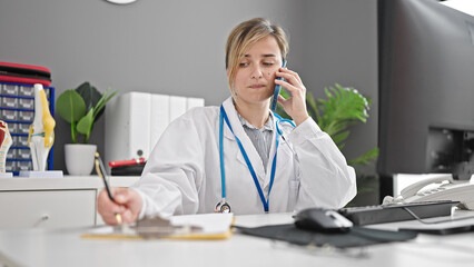 Canvas Print - Young blonde woman doctor talking on smartphone writing on document at clinic