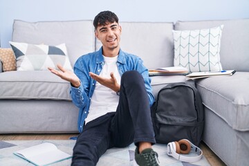 Wall Mural - Young hispanic man sitting on the floor studying for university inviting to enter smiling natural with open hand