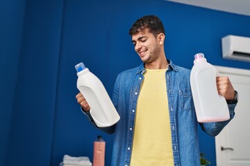 Poster - Young hispanic man washing clothes holding detergent bottles at laundry room