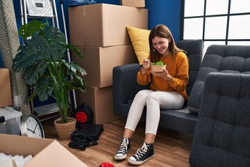 Canvas Print - Young blonde woman eating salad sitting on sofa at new home