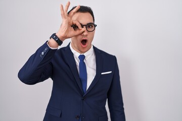 Sticker - Young hispanic man wearing suit and tie doing ok gesture shocked with surprised face, eye looking through fingers. unbelieving expression.