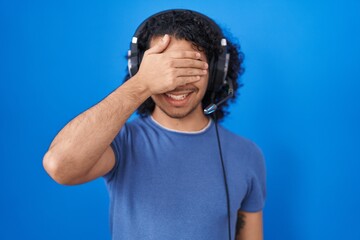 Canvas Print - Hispanic man with curly hair listening to music using headphones smiling and laughing with hand on face covering eyes for surprise. blind concept.