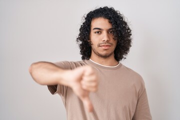 Wall Mural - Hispanic man with curly hair standing over white background looking unhappy and angry showing rejection and negative with thumbs down gesture. bad expression.