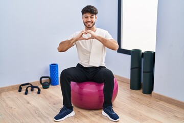 Poster - Hispanic man with beard sitting on pilate balls at yoga room smiling in love doing heart symbol shape with hands. romantic concept.