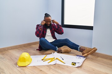 Sticker - African american man sitting on the floor at new home looking at blueprints covering ears with fingers with annoyed expression for the noise of loud music. deaf concept.