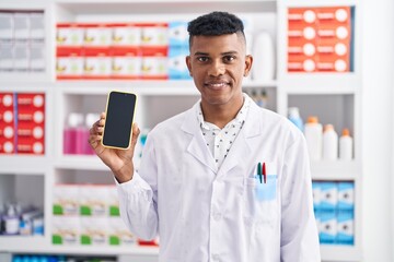 Poster - Young hispanic man working at pharmacy drugstore showing smartphone screen looking positive and happy standing and smiling with a confident smile showing teeth