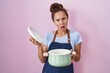 © Krakenimages.com - Brunette woman wearing apron holding cooking pot in shock face, looking skeptical and sarcastic, surprised with open mouth