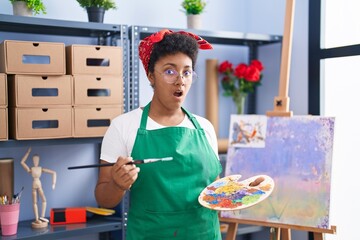 Wall Mural - Young african american woman drawing on canvas at art studio in shock face, looking skeptical and sarcastic, surprised with open mouth