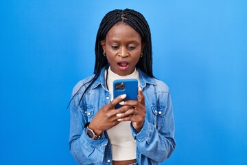 Sticker - African woman with braids using smartphone typing message scared and amazed with open mouth for surprise, disbelief face