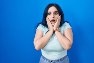 Canvas Print - Young modern girl with blue hair standing over blue background afraid and shocked, surprise and amazed expression with hands on face