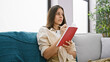 © Krakenimages.com - Attractive young hispanic woman savors tranquil moments of comfort, lounging on her sofa at home. lost in deep thought she pens her ideas, writing in her book amidst the cozy room background.